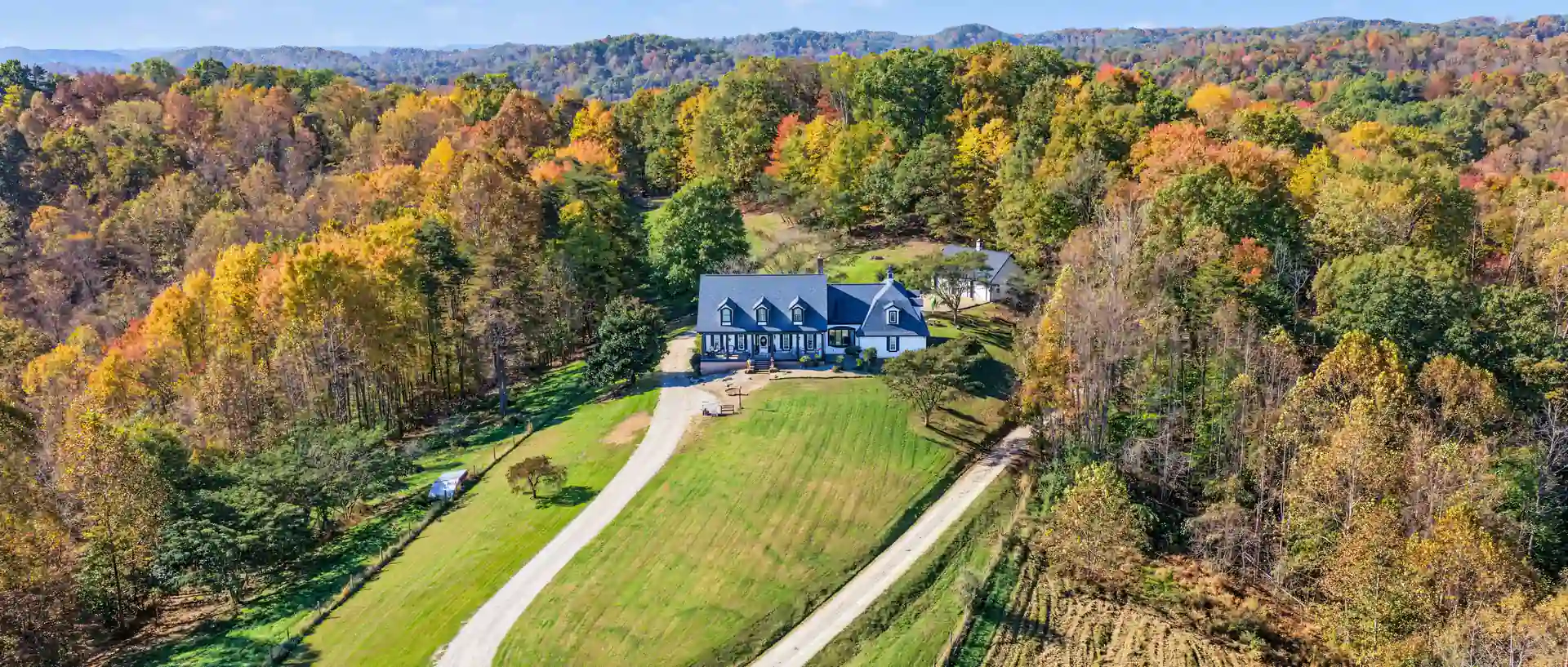 Aerial of the white house at the head of a looping gravel drive, a mown green lawn and a plowed field below, fall-color woods all around. 469 Vincent Hill Rd, Cairo, West Virginia, shot for Whitetail Properties.