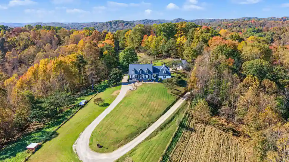 Aerial of the white house at the head of a looping gravel drive, a mown green lawn and a plowed field below, fall-color woods all around. 469 Vincent Hill Rd, Cairo, West Virginia, shot for Whitetail Properties.