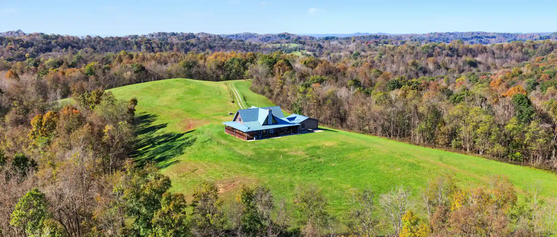 Aerial of the blue-roofed log home on its green hilltop meadow, ringed by fall-color woods, with ridgelines in the distance. Roane County, West Virginia, shot for Whitetail Properties.