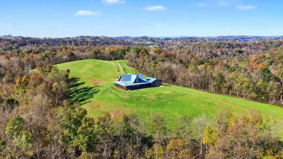 Aerial of the blue-roofed log home on its green hilltop meadow, ringed by fall-color woods, with ridgelines in the distance. Roane County, West Virginia, shot for Whitetail Properties.