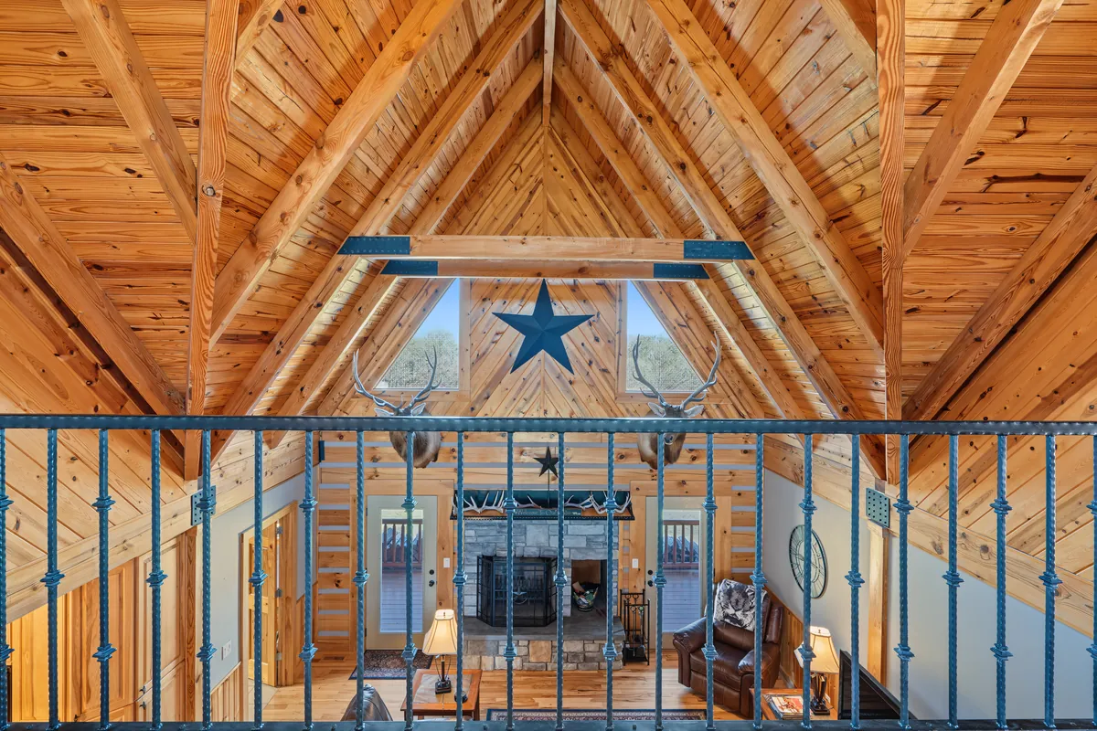 A-frame pine rafters with a metal star and two elk mounts, seen through the loft's iron railing down to the stone fireplace below. Roane County, West Virginia, shot for Whitetail Properties.