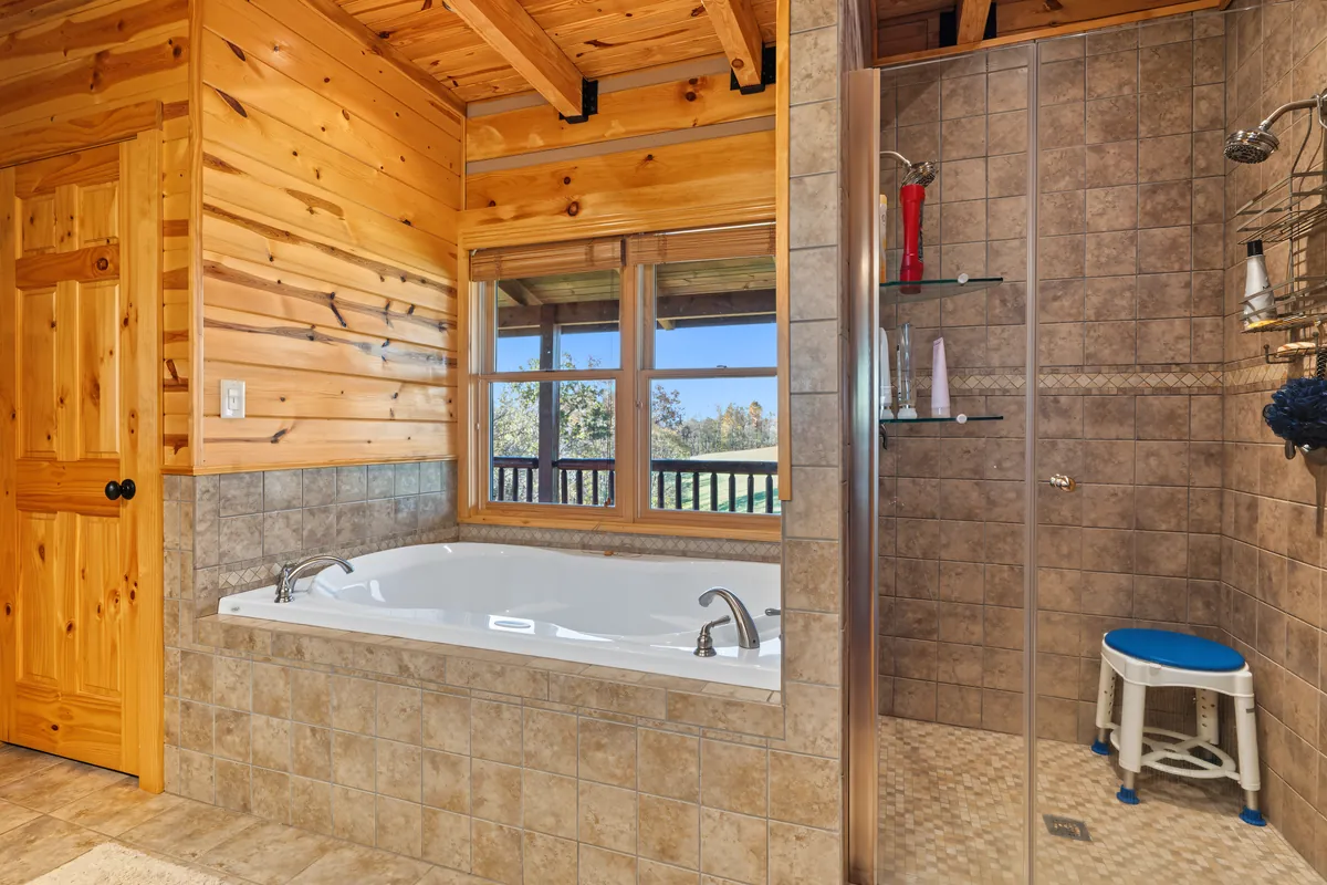 Tiled soaking tub under a window that looks out to the porch rail and the green hilltop, with a walk-in glass shower to the right. Roane County, West Virginia, shot for Whitetail Properties.