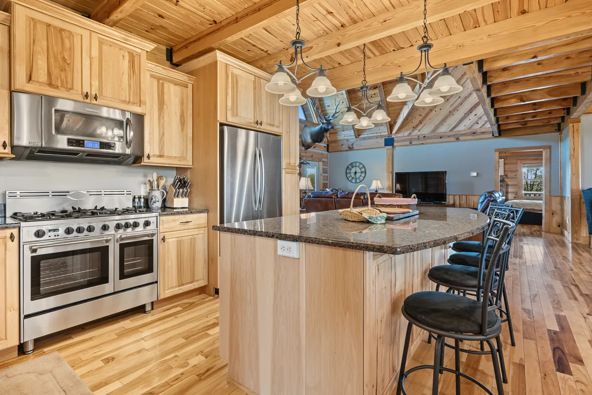 Chef's kitchen with a stainless double-oven range, a stainless refrigerator, a curved granite island with bar stools and the great room behind. Roane County, West Virginia, shot for Whitetail Properties.