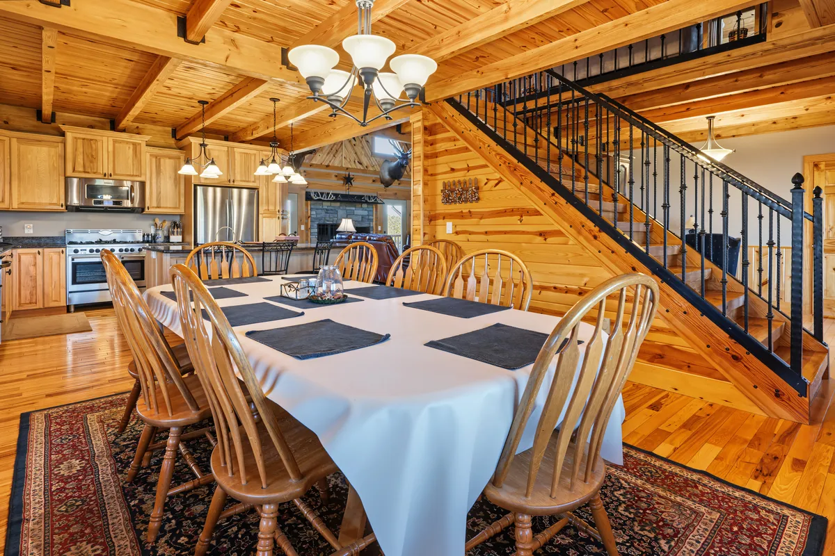Dining table with wooden spindle-back chairs beside the open pine stair with a black iron railing, the kitchen and stone fireplace in the background. Roane County, West Virginia, shot for Whitetail Properties.