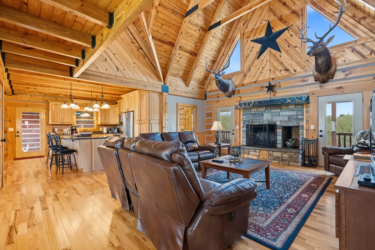 Vaulted pine great room in a log home in Roane County, West Virginia, with a stone fireplace, two elk mounts, brown leather sofas and the kitchen beyond. Shot for Whitetail Properties.