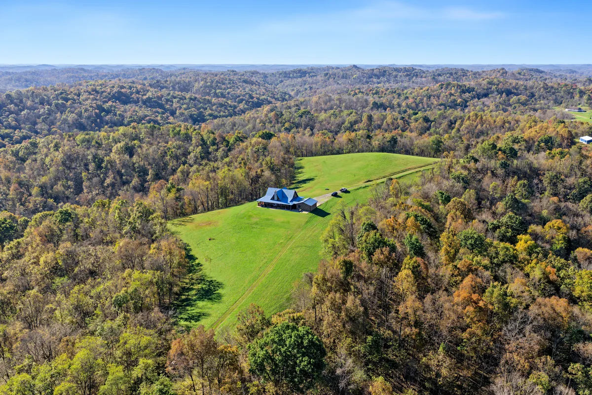 Wide aerial of the log home on its hilltop clearing above rolling hills in fall color, a drive running up through the grass. Roane County, West Virginia, shot for Whitetail Properties.