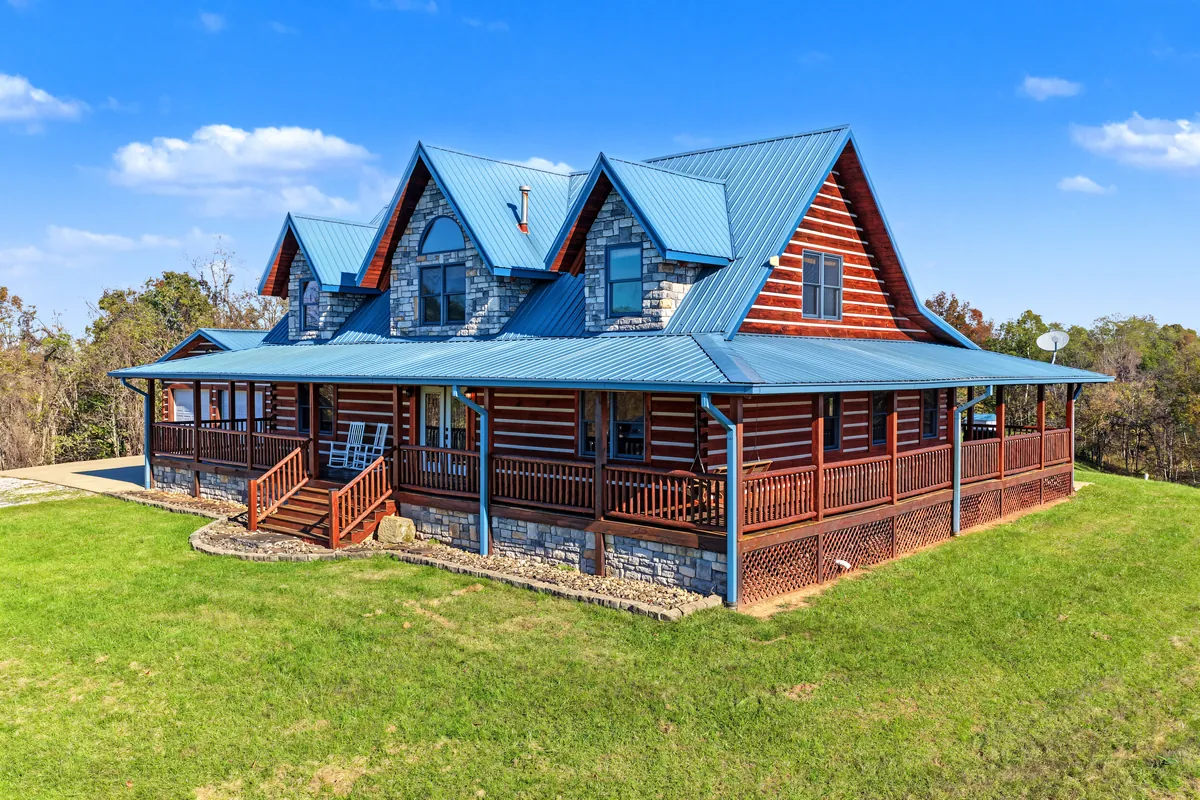 Drone view of a log home with a blue metal roof, stone-faced dormers and a wraparound porch, on a mown green lawn under a blue sky. Roane County, West Virginia, shot for Whitetail Properties.