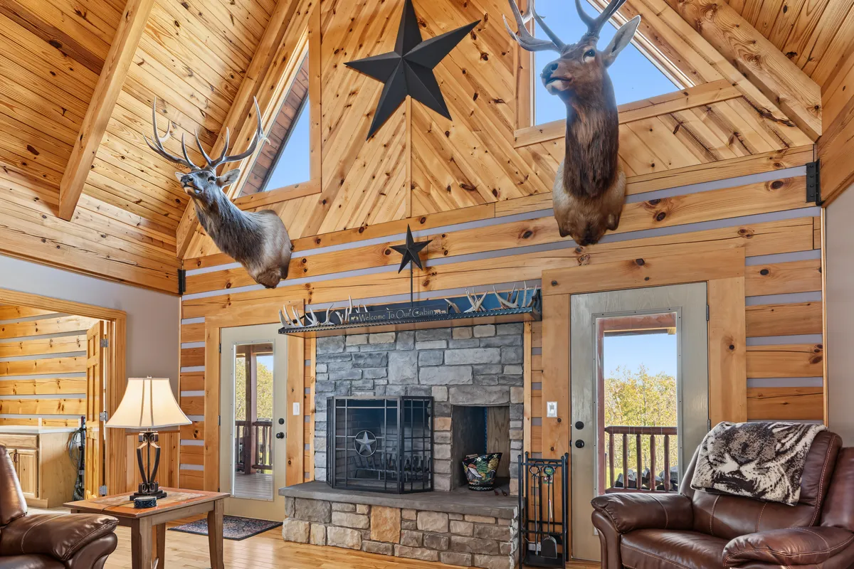 Stone fireplace with a black iron screen and a Texas star under two elk mounts on a pine wall, with a glass-paned door open to the porch on each side. Roane County, West Virginia, shot for Whitetail Properties.