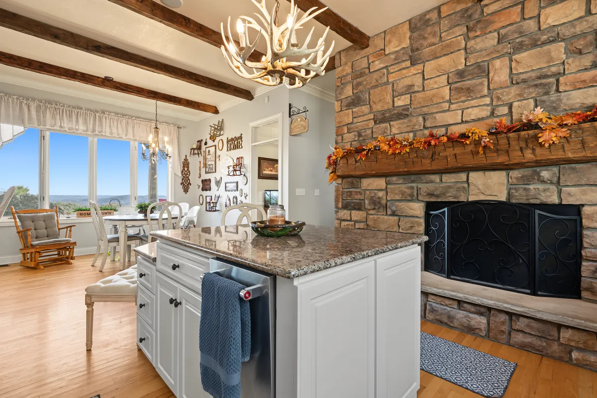 White kitchen island and breakfast area beside a stone fireplace with a fall garland on its beam mantel, windows with a valley view and a hand-hewn beam overhead. 469 Vincent Hill Rd, Cairo, West Virginia, shot for Whitetail Properties.