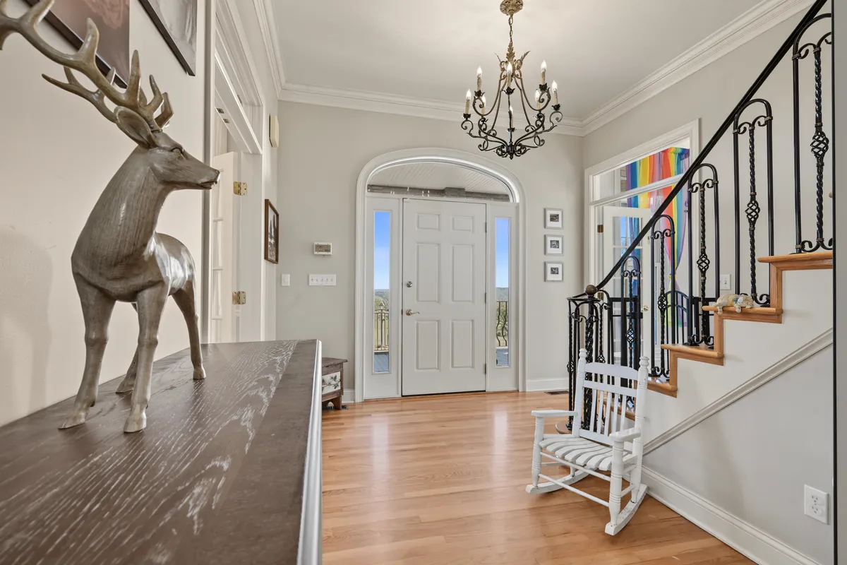 Entry foyer with a wrought-iron chandelier, an arched white front door, a wooden stair with scrolled iron railings and a white rocking chair. 469 Vincent Hill Rd, Cairo, West Virginia, shot for Whitetail Properties.