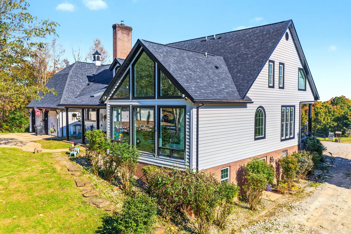 Drone view of a white house with black trim, a tall glass gable wall and a brick chimney, fall-color trees on both sides. 469 Vincent Hill Rd, Cairo, West Virginia, shot for Whitetail Properties.