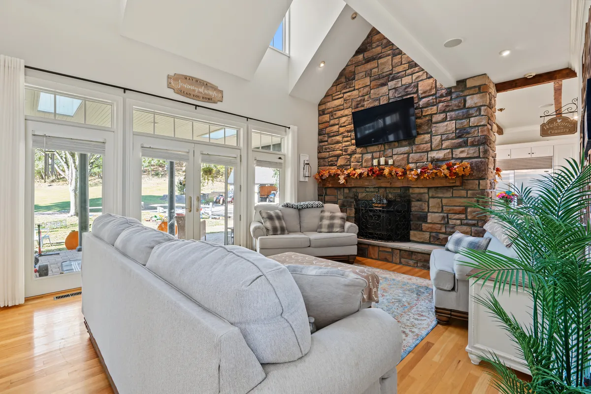 Vaulted living room with a two-story stone fireplace and a mounted television, French doors to the back lawn and light gray sofas. 469 Vincent Hill Rd, Cairo, West Virginia, shot for Whitetail Properties.