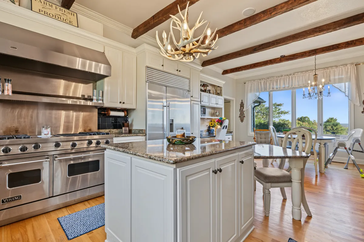 Kitchen with a stainless double-oven range under a hood, white cabinets, a granite island, an antler chandelier and a breakfast nook with a long view out the windows. 469 Vincent Hill Rd, Cairo, West Virginia, shot for Whitetail Properties.