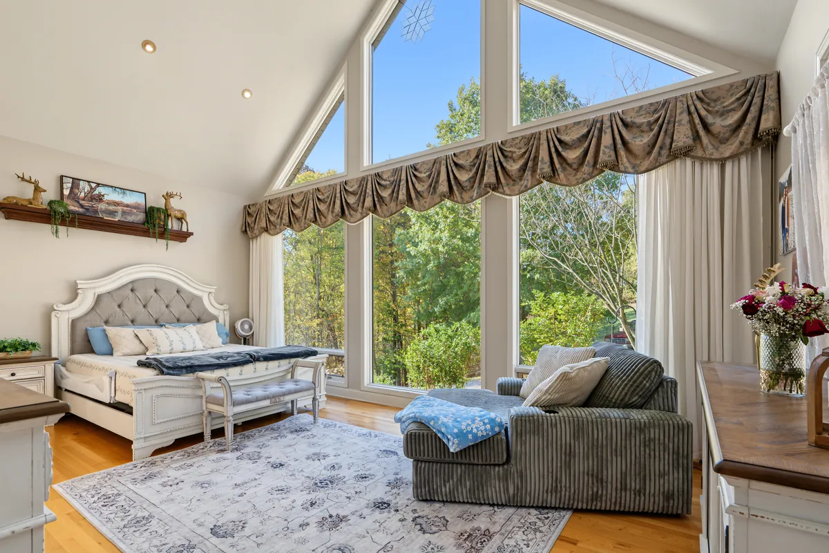 Primary bedroom with a vaulted ceiling and a wall of windows onto the trees, a tufted white bed, a sitting chaise and a patterned rug. 469 Vincent Hill Rd, Cairo, West Virginia, shot for Whitetail Properties.